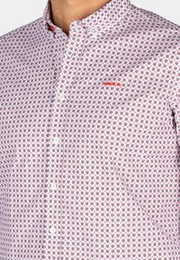 Witte button-up shirt met een rood en zwart bloemenpatroon. Kleine kraag met rode accentstiksels en witte knopen.