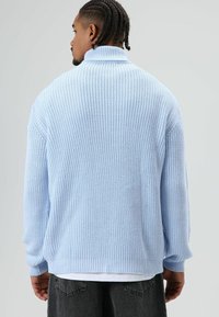 Pull tricoté bleu clair avec un col montant et une texture côtelée. Comprend des manches longues et une coupe décontractée, porté par-dessus un t-shirt blanc.