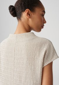 Light beige textured top with a high collar and short sleeves, featuring a subtle striped pattern and a soft woven material.