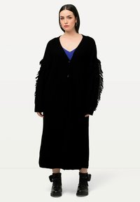 Cardigan nero a maglia con maniche lunghe e frange, aperto davanti e lunghezza a metà polpaccio, indossato sopra una maglietta blu, abbinato a stivali neri.