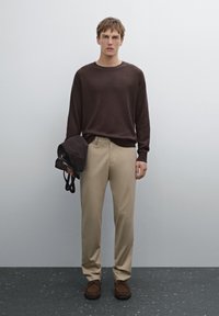 Brown knit sweater, beige tailored trousers, holding a brown bag. Brown shoes with a textured surface. Gray backdrop with subtle shadow.