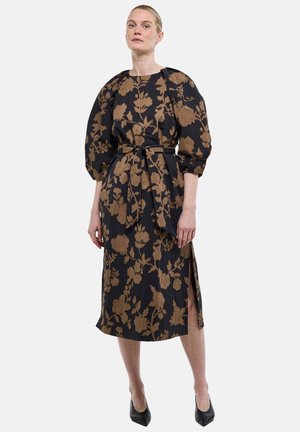 Femme debout portant une robe midi noire à motif floral marron, manches bouffantes, ceinture nouée à la taille, fente latérale et chaussures noires pointues.
