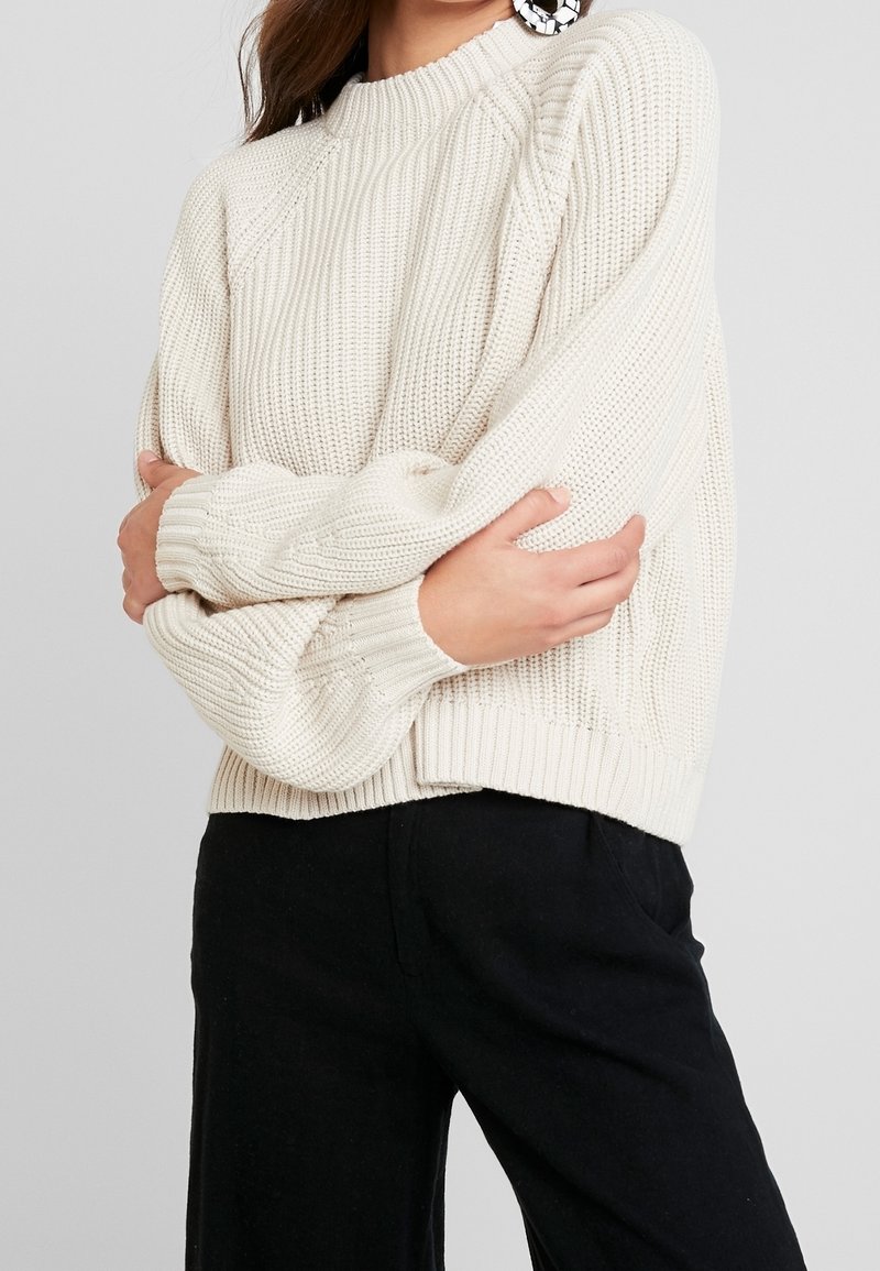 Pull en maille crème avec un col rond et un ourlet côtelé, arborant un motif texturé et une coupe décontractée, associé à un pantalon noir.