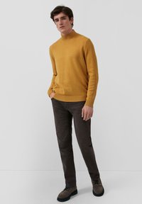 Muž v horčicovožltom roláku, tmavohnedých nohaviciach a hnedých šnurovacích čižmách, stojí s jednou rukou v vrecku.