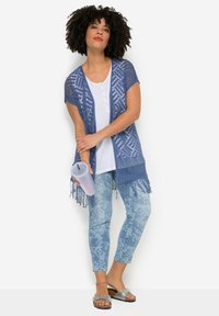 Cardigan blu in maglia aperta con motivi geometrici e orlo frangiato, indossato sopra una maglietta bianca e abbinato a jeans chiari con motivo blu e sandali.