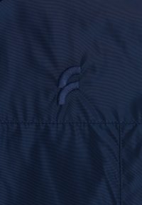Tessuto della giacca blu navy con una texture liscia, caratterizzata da due dettagli ricamati curvi. La cucitura lungo il fondo è ordinata e sicura.