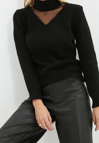 Maglione nero a coste con scollo a V e colletto alto, abbinato a pantaloni di pelle nera. La texture sembra calda e spessa, creando un aspetto accogliente.