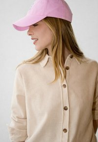 Jeune femme aux longs cheveux blonds portant une chemise en velours côtelé beige clair et une casquette de baseball rose, regardant sur le côté avec un léger sourire.