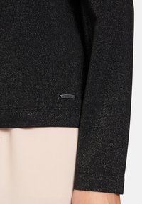 Schwarzer Pullover mit glitzernder Textur, ausgestattet mit einem metallischen Logo am Saum. Lange Ärmel und eine taillierte Passform.