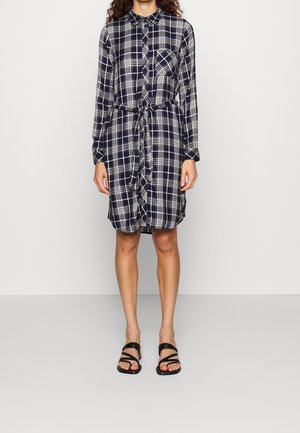 Femme portant une robe-chemise à manches longues à carreaux bleu marine et blanc, avec une poche avant et une ceinture, associée à des sandales noires à brides.