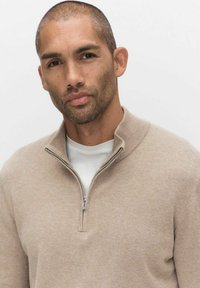 Beżowy sweter zapinany na zamek, z teksturowanym splotem, wyposażony w wysoki kołnierz i srebrny zamek, noszony na białej koszulce. Gładkie tło.