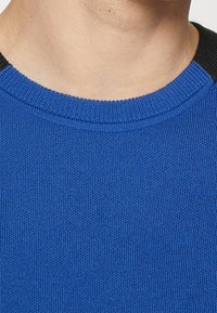 Maglione blu lavorato a maglia con una texture a coste e accenti neri sulle spalle. Presenta un collo rotondo e un design aderente.