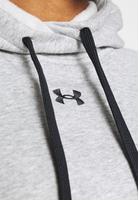 Sudadera gris de tejido suave, con un logo negro bordado y cordones oscuros. Vista en primer plano que enfatiza la textura y el diseño.