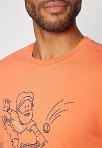 Lähellä kuva miehestä, joka pitää yllään oranssia paitaa, jonka rinnassa on tulostettuna sarjakuvamaailman tennispelaaja, joka lyö palloa.