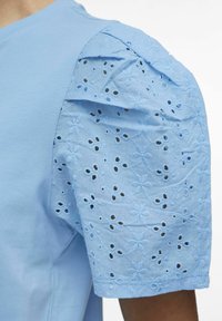 Jasnoniebieska bluzka z gładkim fasonem i bufiastymi rękawami z misternym haftem w kwiatowe oczka oraz wycięciami.