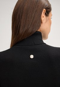 Zwarte ribbel turtleneck met een aansluitend ontwerp, voorzien van een zilveren metalen logo-accent op de achterkant. Zachte textuur en monochromatische kleurenschema.