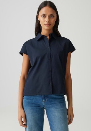 Vrouw met lang donker haar, een marineblauw kortermig overhemdd met knopen en blauwe spijkerbroek tegen een eenvoudige achtergrond.