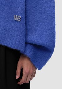 Blauer gestrickter Pullover mit lockerem Design und flauschiger Textur. Verfügt über ein kleines silbernes WB-Logo am Ärmel. Die Hand liegt in der Nähe des Saums.