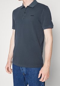 Koszulka polo w ciemnoszarym kolorze, z klasycznym kołnierzykiem, krótkimi rękawami oraz strukturalnym materiałem. Czarny logo na lewej piersi. Noszona z niebieskimi dżinsami.