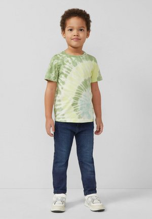 Jeune garçon debout, portant un t-shirt tie-dye vert et jaune, un jean bleu foncé et des baskets blanches, sur un fond gris uni.