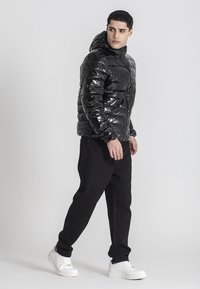 Giovane uomo che cammina, indossando un giubbotto a vento nero lucido con cappuccio, pantaloni neri e sneaker bianche, che guarda oltre la spalla.