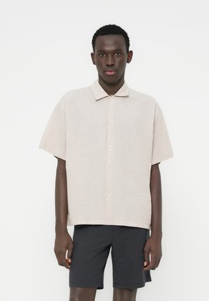 Κοντομάνικο, ανοιχτό μπεζ πουκάμισο με κουμπιά, κατασκευασμένο από μαλακό ύφασμα με γιακά και άνετη εφαρμογή, σε συνδυασμό με σκούρο σορτς.