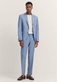 Hellblauer Anzug mit Zweiknopfjacke und schmal geschnittenen Hosen. Maßgeschneiderte Passform, strukturiertem Stoff, kombiniert mit einem weißen Rundhals-Shirt und braunen Schuhen.