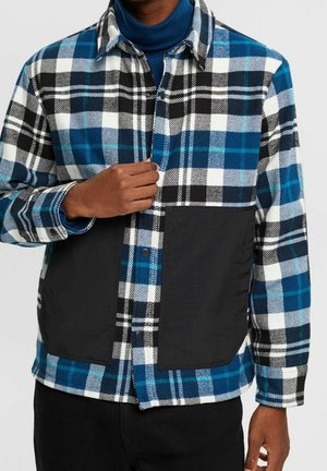 Homme portant une chemise à carreaux bleue, noire et blanche avec de grandes poches avant noires, boutonnant la chemise sur un col roulé bleu.