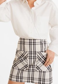 Chemise blanche à boutons avec une poche, associée à une mini-jupe écossaise beige ornée de motifs à carreaux noirs et gris, dotée de deux poches avant.
