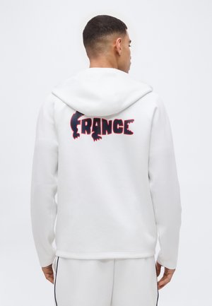 Hombre con sudadera con capucha blanca con un logo "FRANCIA" en azul oscuro y rojo en la espalda, de pie frente a un fondo blanco liso.
