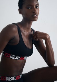 Zwarte sportbeha met een brede witte en rode band met het "DIESEL" logo. Het model toont een natuurlijke huidtextuur en een zelfverzekerde pose.