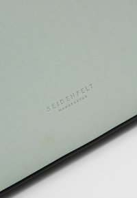 Jasnozielona teksturowana skórzana powierzchnia z wytłoczonym logo "Seidenfelt Manufaktur" w pobliżu dolnej krawędzi.