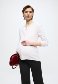 Hellrosa, knöpfbare Strickjacke mit rundem Ausschnitt und dreiviertellangen Ärmeln, kombiniert mit schwarzer Hose und einer kleinen roten Handtasche.