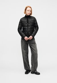 Γυναίκα με κοντά κόκκινα μαλλιά, ντυμένη με μαύρο μπουφάν με επένδυση, ξεθωριασμένο γκρι τζιν με φαρδιά μπατζάκια και μαύρα γυαλιστερά μοκασίνια, στέκεται σε λευκό φόντο.