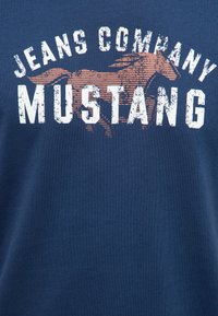 Tissu bleu marine avec texte blanc usé "JEANS COMPANY MUSTANG" et un cheval stylisé en train de courir marron derrière le texte.