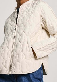 Prošívaná krémová bunda se zapínáním na zip vpředu, kulatým límcem a bočními kapsami. Má jemný diamantový vzor a štítek s nápisem "Pepe Jeans."