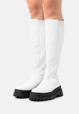 Bottes blanches montantes jusqu'aux genoux avec semelles épaisses et noires portées sur des jambes nues sur un fond clair.
