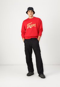 Felpa rossa con logo di coccodrillo giallo, abbinata a pantaloni cargo neri e sneakers scure, indossata con un cappello a secchiello nero.