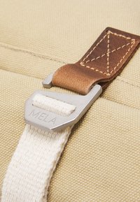 Tan canvas tas met een zilveren metalen sluiting. De band is van crème-kleurige katoen en een donkerbruine leren accent is zichtbaar op de sluiting.