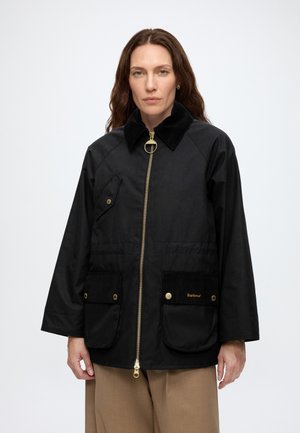 Femme portant une veste Barbour noire avec une fermeture éclair dorée, des poches à boutons, un col noir et un pantalon beige sur un fond uni.
