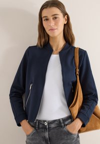 Jeune femme portant une veste zippée bleu marine sur un haut blanc, jean gris, avec un sac à bandoulière en daim marron, debout devant un mur clair uni.