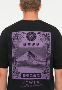 Man wearing black shirt with purple graphic of Mount Fuji, floral border, Japanese characters, and text "FROM JAPAN WITH LOVE" on the back.