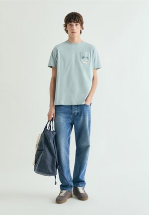 Joven con camiseta gráfica azul claro, vaqueros azules y zapatillas deportivas marrones, sosteniendo una mochila azul marino con una mano.