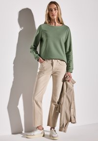 Zelená mikina, béžové zkrácené kalhoty s dekorativními kapsami a bílé tenisky. Béžová bunda je přehozena přes ruku.