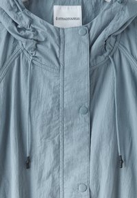 Veste à capuche bleu clair avec fermeture devant à boutons-pression et cordons ajustables, étiquetée avec la marque Stradivarius à l’intérieur du col.
