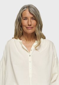 Blusa di un leggero color crema realizzata in un tessuto morbido e testurizzato. Presenta una chiusura frontale a bottoni, un collo leggermente arricciato e maniche larghe e rilassate.