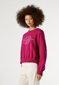 Žena s kudrnatými vlasy, oblečená v magentovém svetru s bílým textem ve tvaru rtů a krémových kalhotách, stojící na jednobarevném světlém pozadí.