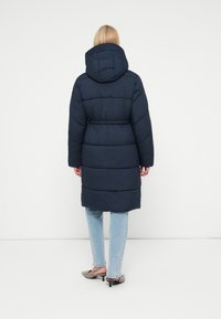 Geacă puffer bleumarin cu glugă, având talie strânsă și un model matlasat orizontal. Pur tată peste blugi bleu deschis și pantofi cu toc.