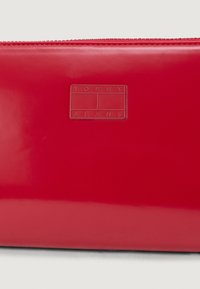 Rød blank lommebok med glidelås og preget "TOMMY JEANS"-logo. Glatt tekstur og rektangulær form.