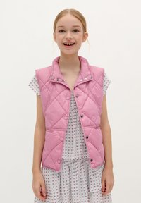Roze gewatteerde vest met een hoge kraag, drukknoopsluiting en twee zijzakken, gedragen over een witte jurk met een patroon en korte mouwen.